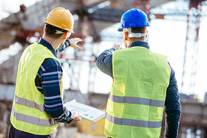Two men in hardhats and safety vests at jobsite with graphs in hand pointing at site location.