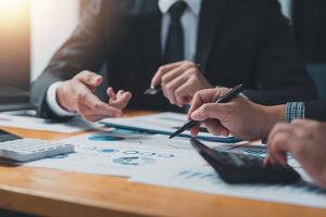 Two men in suits holding pens and going over graphs on papers with calculators in an office.