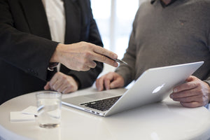 Consulant in suit pointing at macbook with a pen in hand and a glass of water on the table.