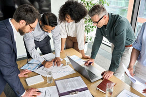 Team standing in circle over table going through paper for project in an office with glasses of water.