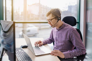 Technology consultant working remotely with headset and laptop next to monitor in an office.